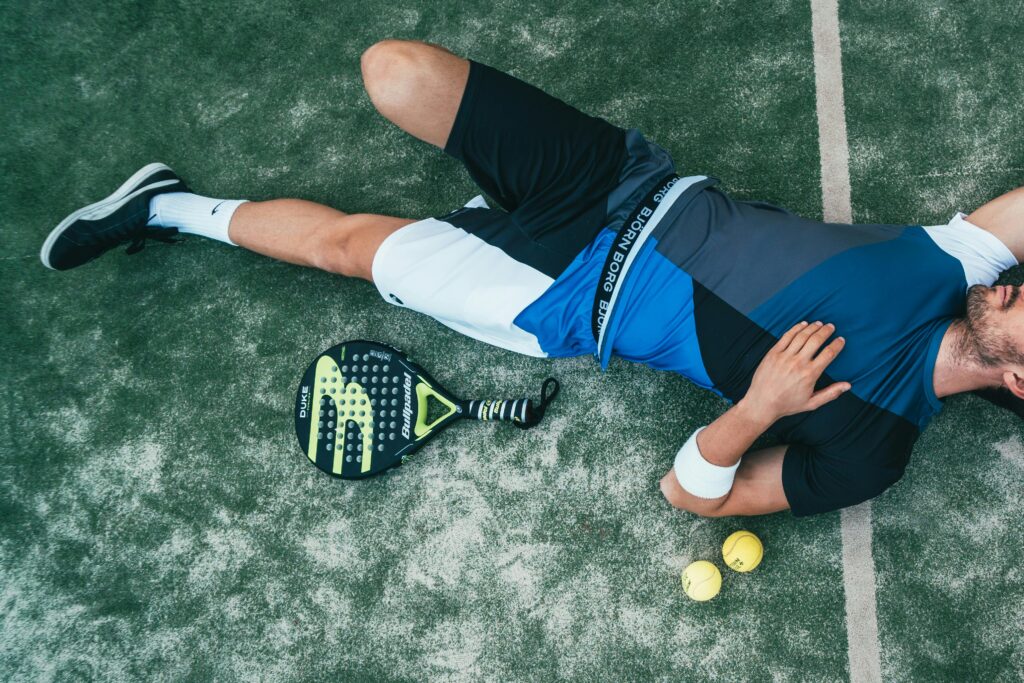 Tired young athlete resting on padel court with racket and balls nearby.